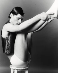 Black and white portrait of a male model wearing a dark leather vest and white underwear, seated on a stool.