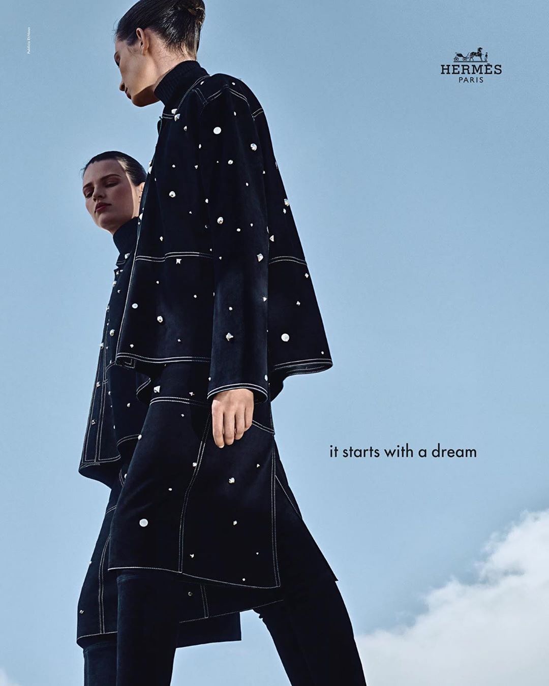 Two women in matching dark coats with pearl embellishments and black pants pose against a blue sky in a fashion editorial shot.