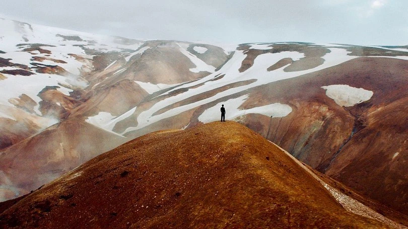 A lone adult stands on a red-brown hilltop overlooking an expansive landscape of snow-patched mountains under a grey sky.