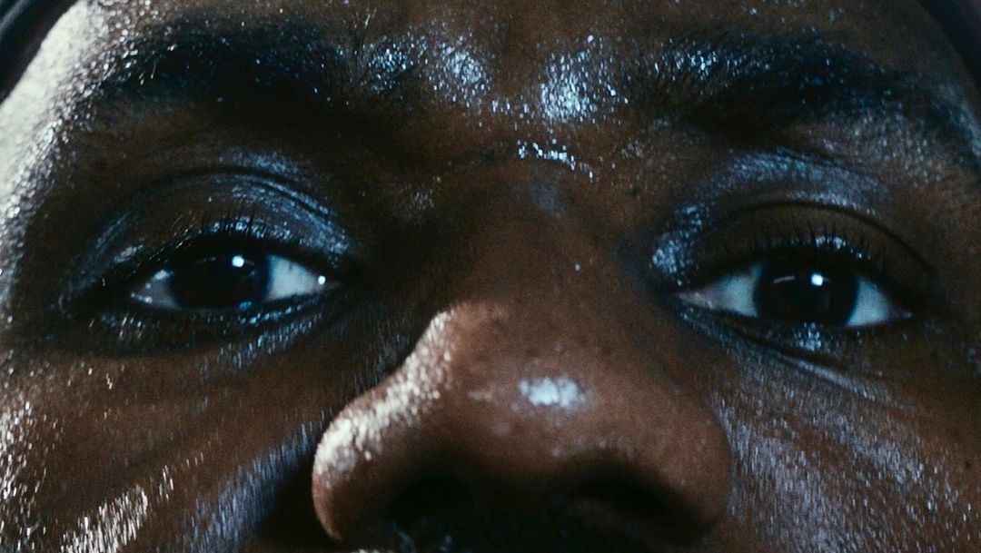 Macro close-up of a sweaty, dark-skinned adult's face, highlighting the eyes, nose, and forehead, set against a dark background.