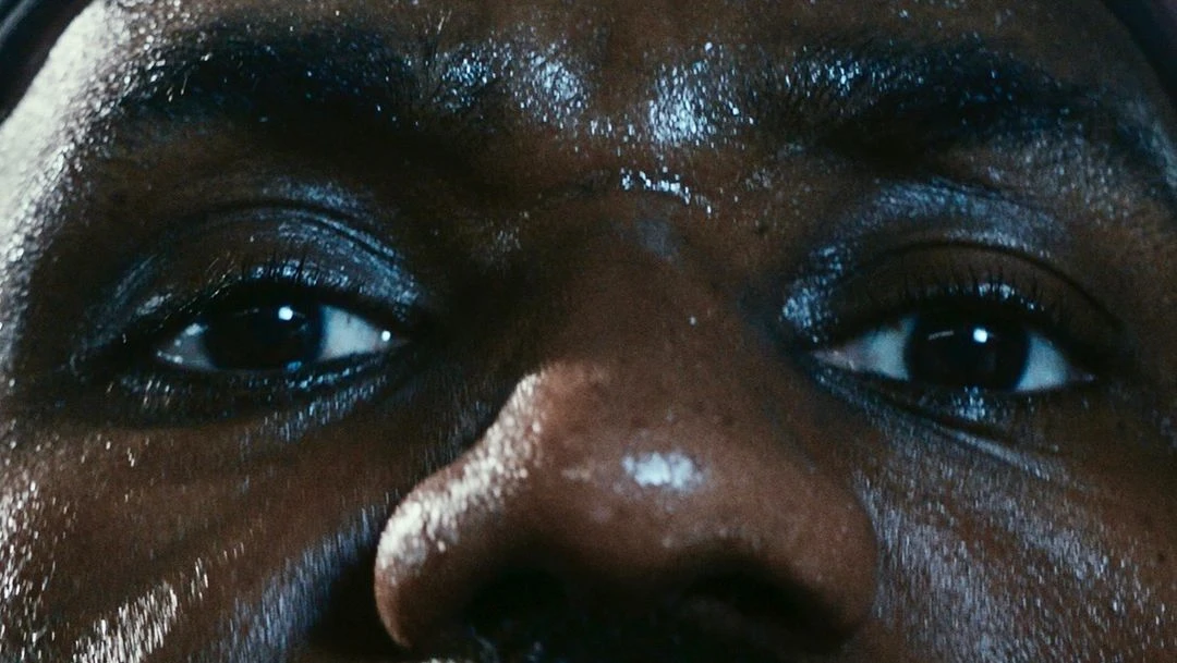 Macro close-up of a sweaty, dark-skinned adult's face, highlighting the eyes, nose, and forehead, set against a dark background.