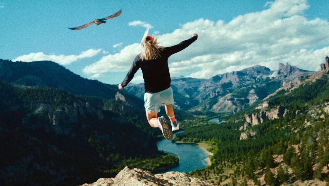 A person jumps off a rock cliff with arms outstretched; mountains, trees, and a river create a stunning backdrop with a bird in flight.