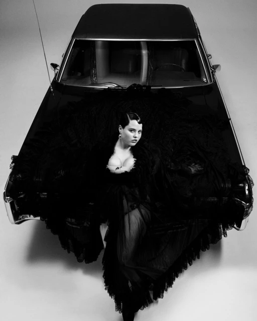 Black and white portrait of a woman with short dark hair laying on the hood of a car wearing a sheer tulle dress.