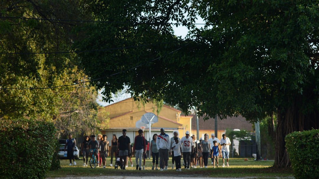 A group of people of varied ages walking on a path in an outdoor neighborhood, lined with trees and homes during the daytime.