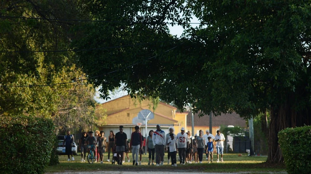 A group of people of varied ages walking on a path in an outdoor neighborhood, lined with trees and homes during the daytime.