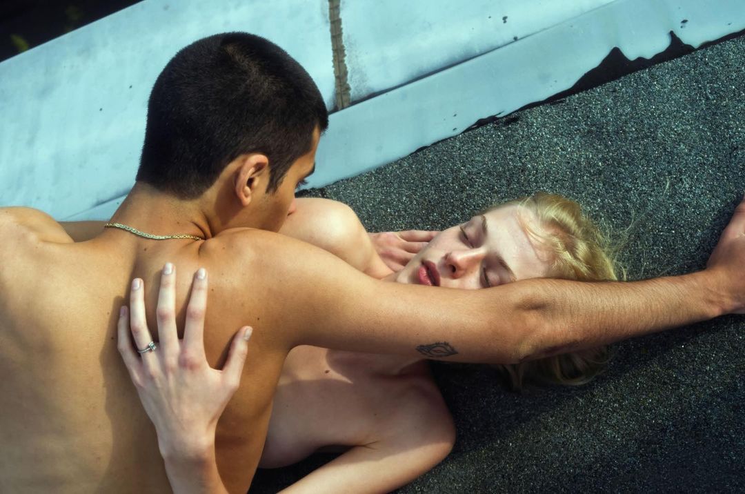 Intimate portrait of a shirtless couple laying on a textured surface, one with dark hair and gold chain, the other blonde and fair-skinned.