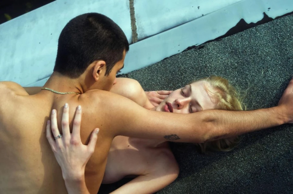 Intimate portrait of a shirtless couple laying on a textured surface, one with dark hair and gold chain, the other blonde and fair-skinned.