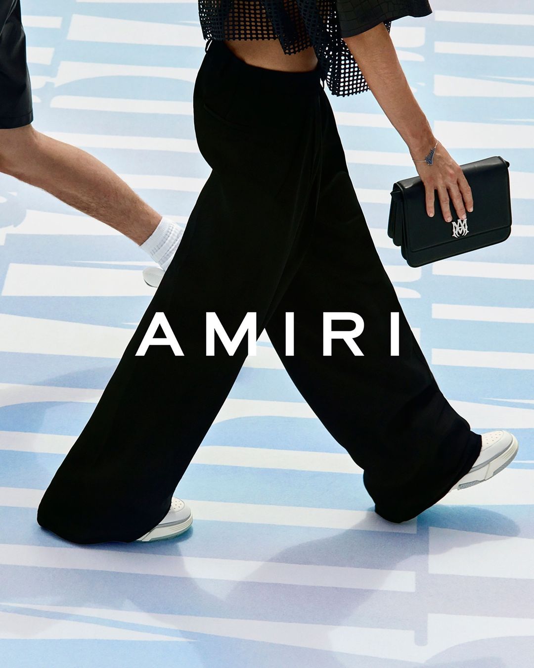 Fashion shot featuring models walking, showcasing black pants, a black handbag, and white sneakers on a light blue background. 