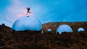A person jumps on a silver sphere in a desert landscape against a blue sky with a distant canyon backdrop. Action shot.