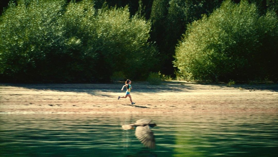 Woman running on beach near lake and forest, reflected in water. Outdoor fitness, nature, and sport concept with greenery and sunlight.