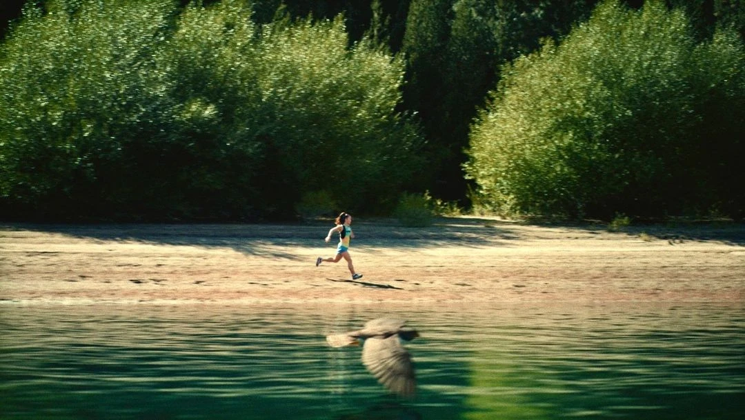 Woman running on beach near lake and forest, reflected in water. Outdoor fitness, nature, and sport concept with greenery and sunlight.