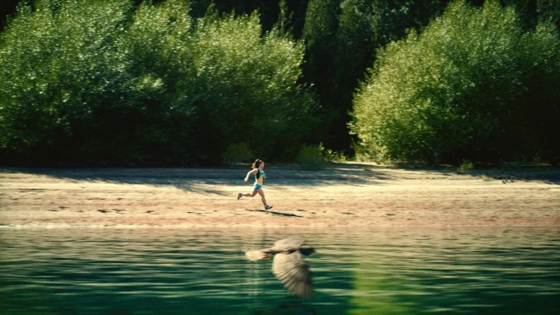 Woman running on beach near lake and forest, reflected in water. Outdoor fitness, nature, and sport concept with greenery and sunlight.
