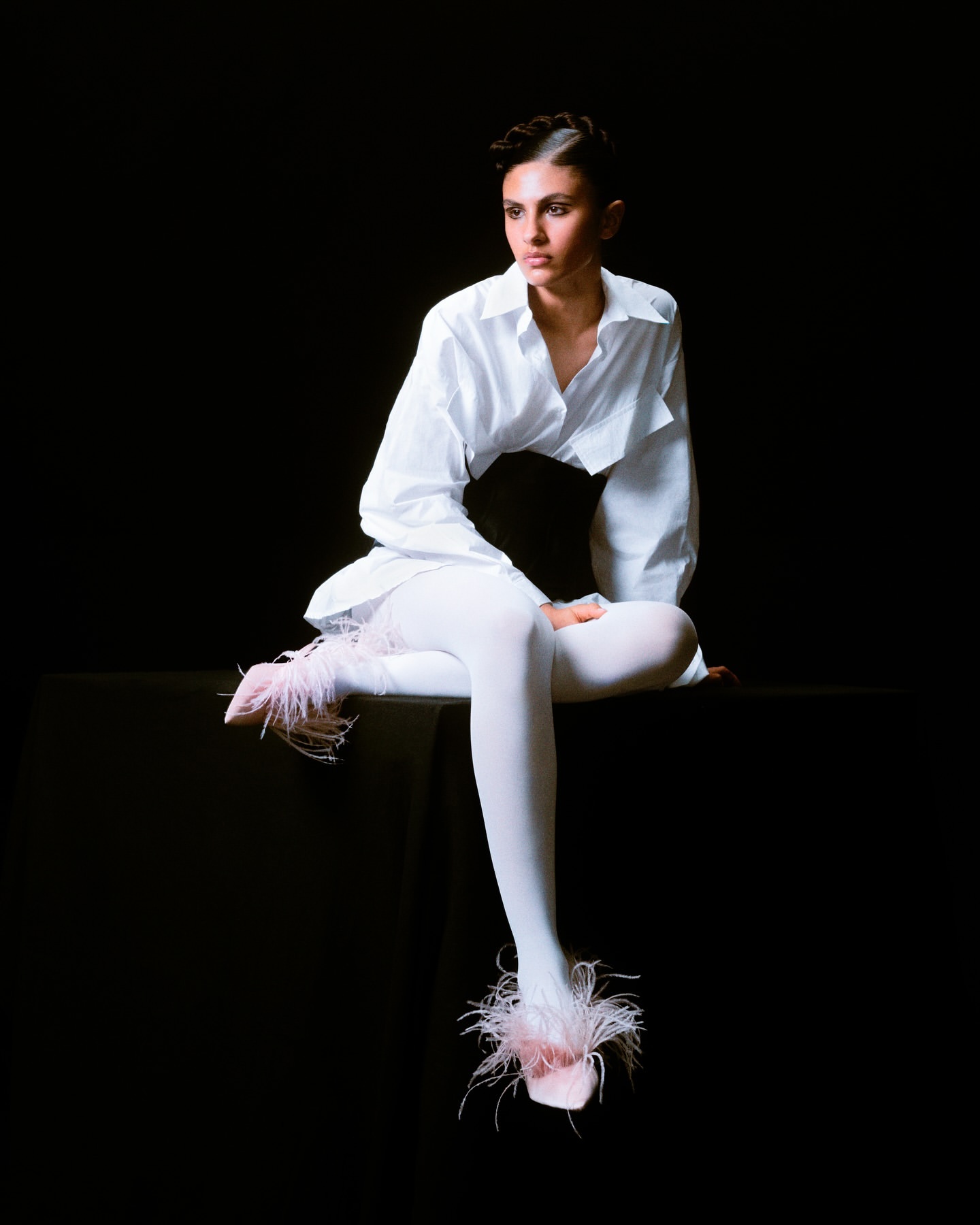 Elegant woman in white shirt and tights, seated on a black surface, strikes a pose against a dark backdrop with stylish feathered footwear.