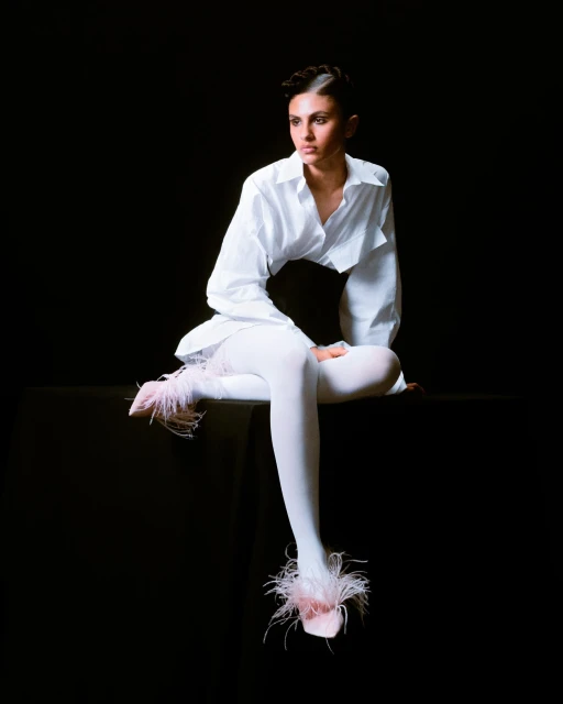Elegant woman in white shirt and tights, seated on a black surface, strikes a pose against a dark backdrop with stylish feathered footwear.