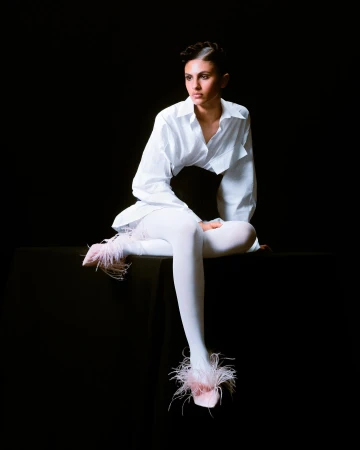 Elegant woman in white shirt and tights, seated on a black surface, strikes a pose against a dark backdrop with stylish feathered footwear.