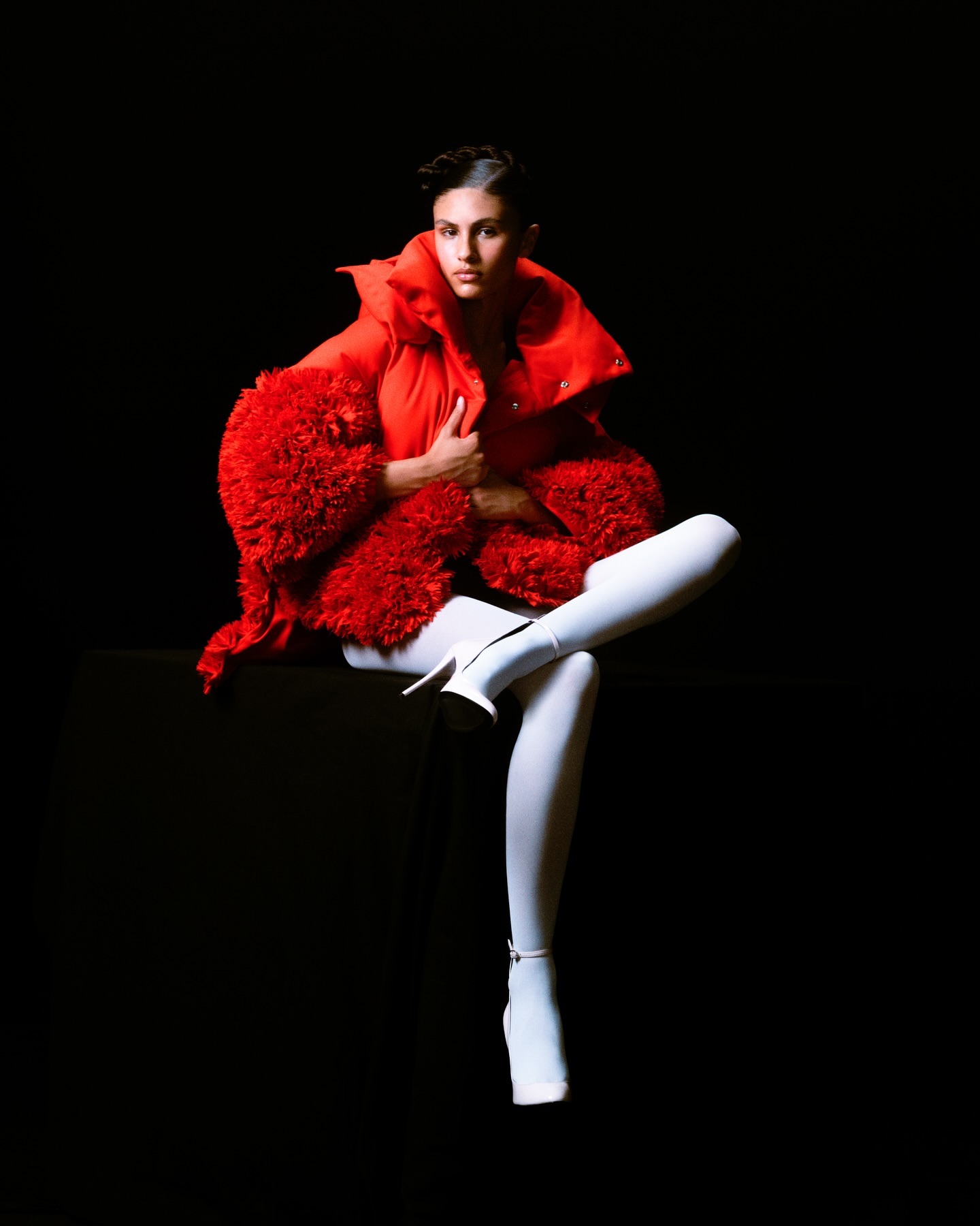 Woman in red puffer jacket and white leggings sits against a black backdrop, creating a striking and fashionable high-contrast visual.