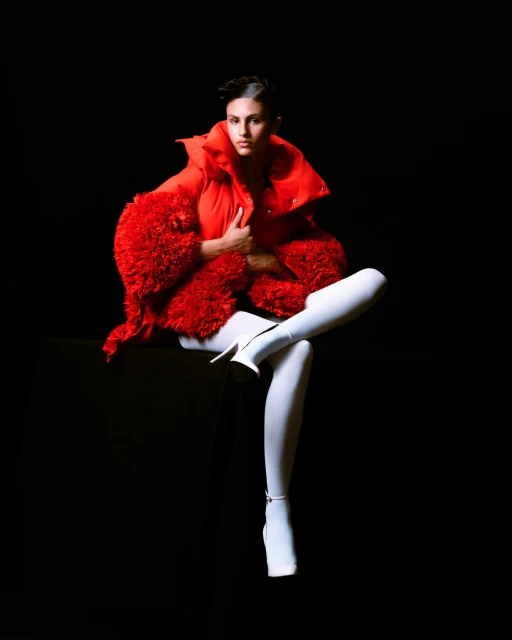Woman in red puffer jacket and white leggings sits against a black backdrop, creating a striking and fashionable high-contrast visual.