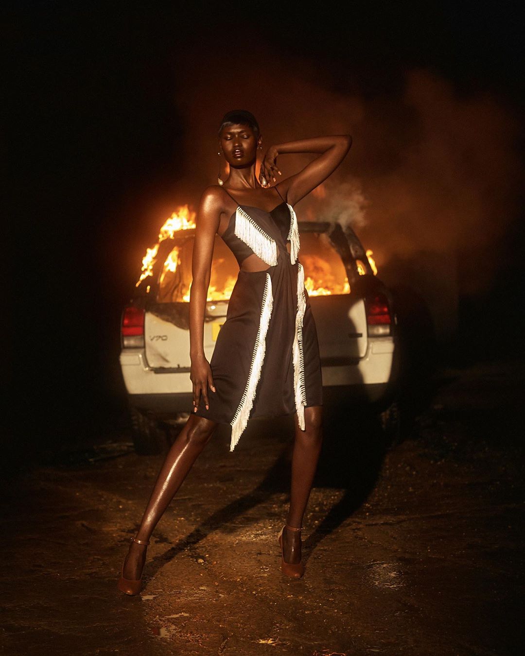 An adult female model poses in a stylish black dress with white fringe, with a burning car in the background on a dark night.