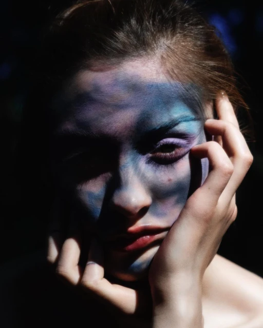 Close-up portrait of a woman with dramatic blue and purple face paint in dark, moody lighting, with a hand near her face.