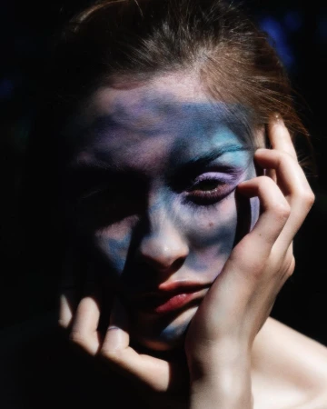 Close-up portrait of a woman with dramatic blue and purple face paint in dark, moody lighting, with a hand near her face.