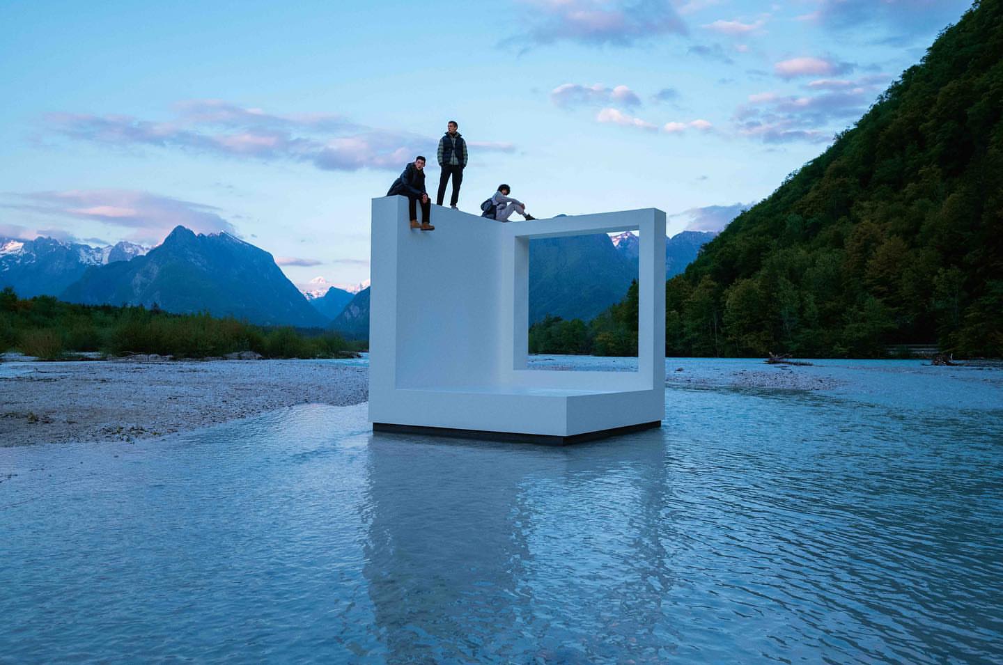 Three people pose atop a modern white geometric structure in a river, set against a background of mountains and a tranquil sky.