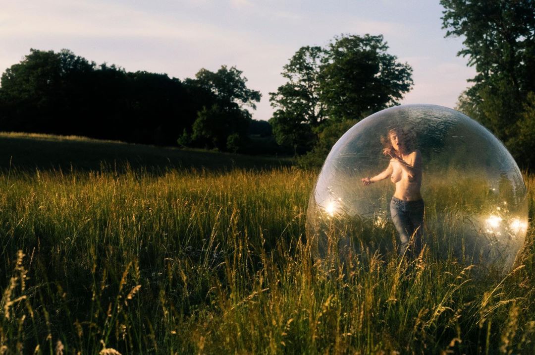 A woman stands inside a large transparent bubble in a field of tall grass. The scene is outdoors during golden hour with trees in the background.