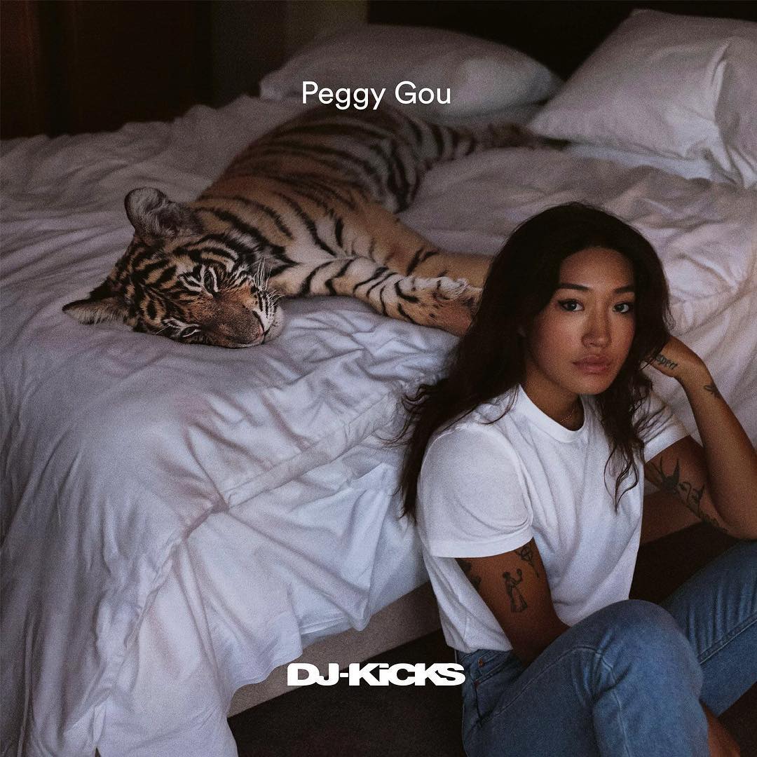 Portrait of a woman in a white t-shirt and blue jeans sitting on a bed next to a tiger plush toy in a bedroom setting.