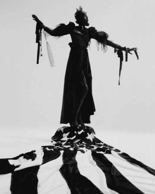 Monochrome full shot of a person in a dark, elaborate gown and headpiece standing on a fabric with stars and stripes pattern.