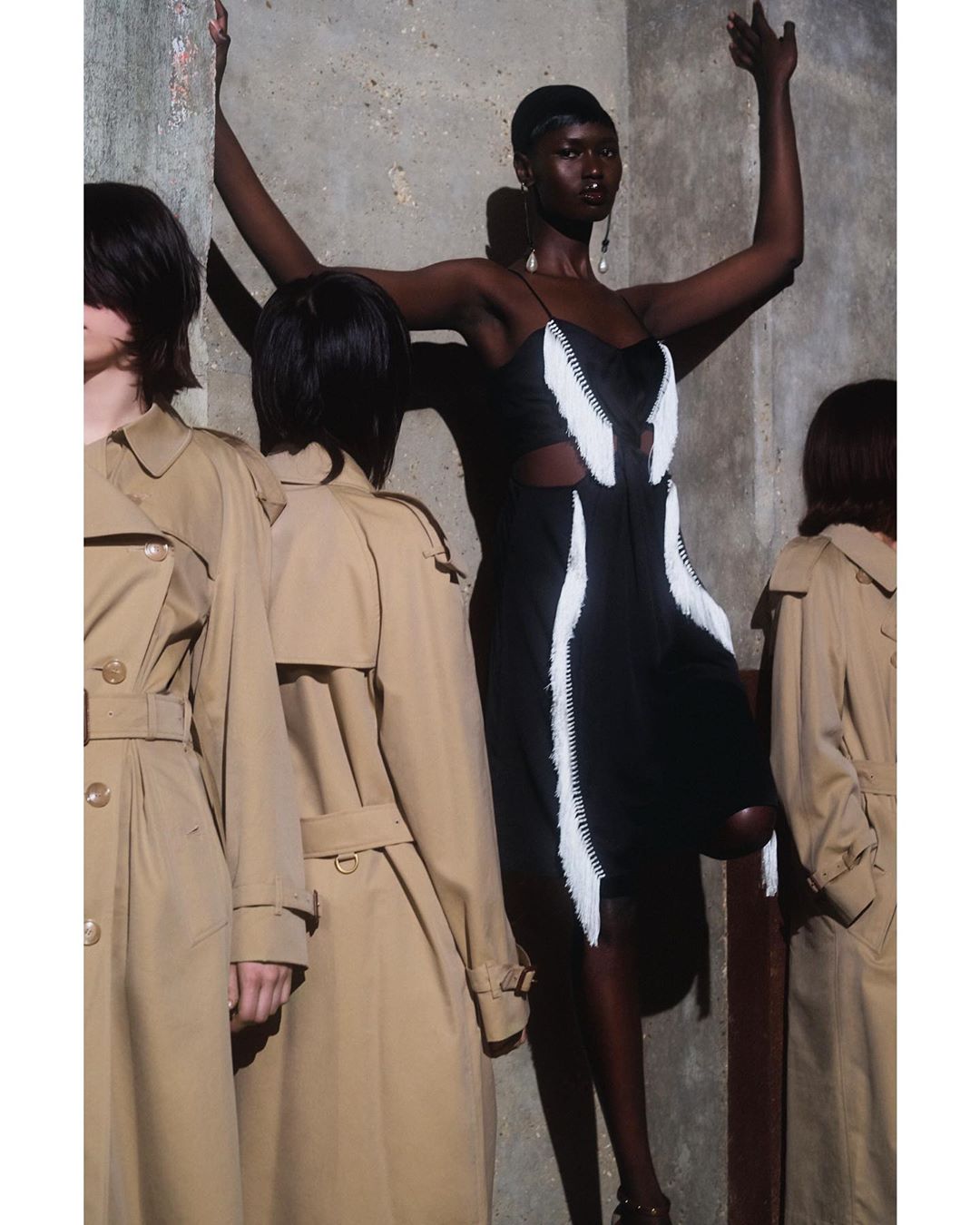 Fashion editorial shot of a Black woman in a black and white dress with other women in trench coats in the background against concrete walls.