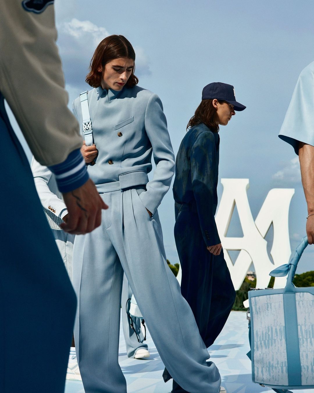 Fashion editorial image showcasing adult male models in light blue suits and caps in an outdoor setting with a clear sky.