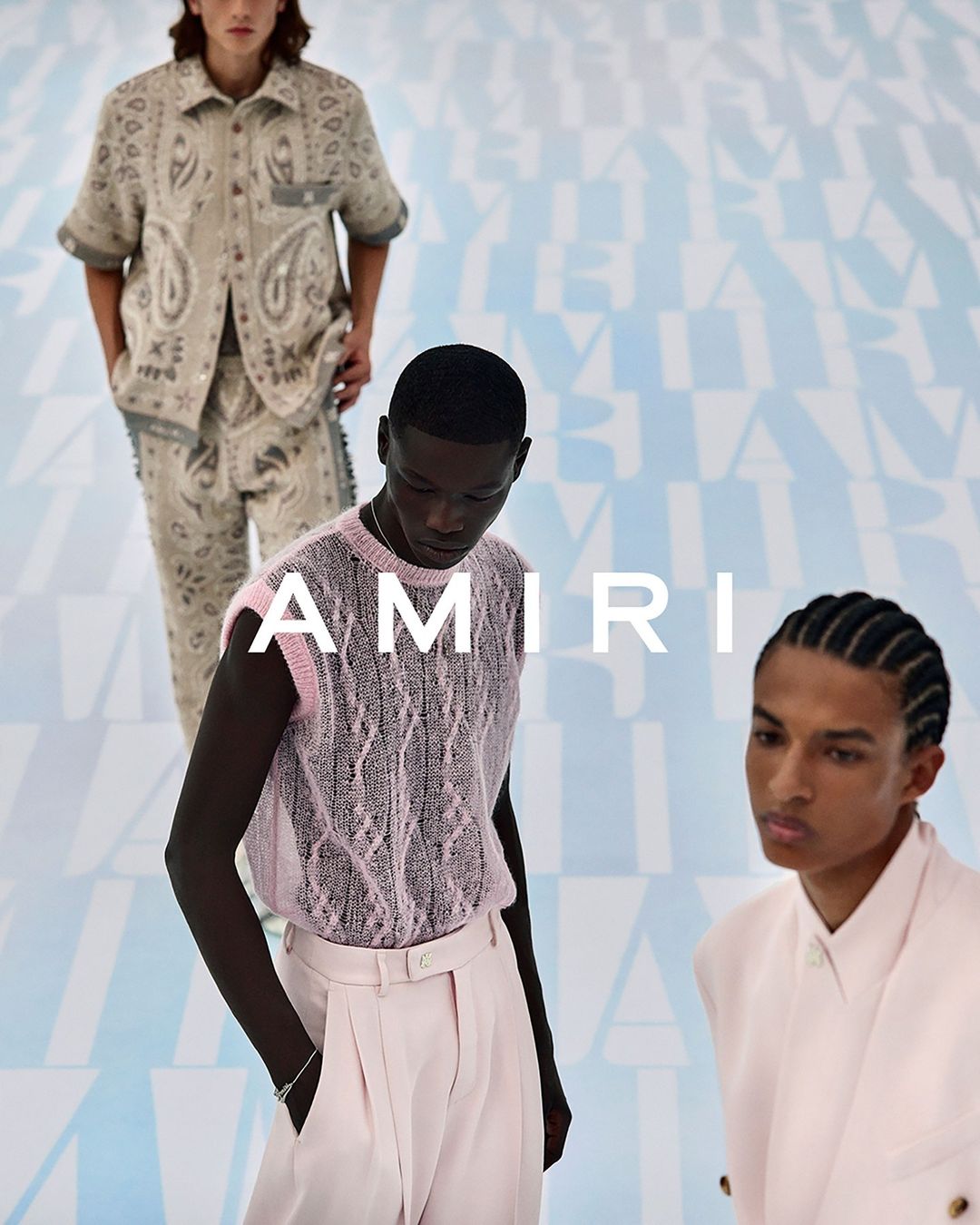 Three dark and light-skinned male fashion models showcase AMIRI clothing, including paisley patterns and knitwear, against a branded backdrop.