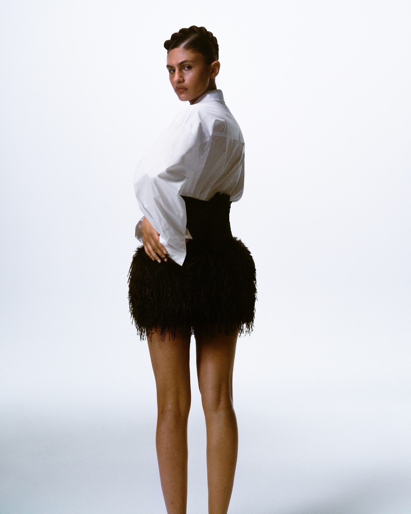 Model with braided hair wearing a white button-down shirt and black feathered skirt against a white background