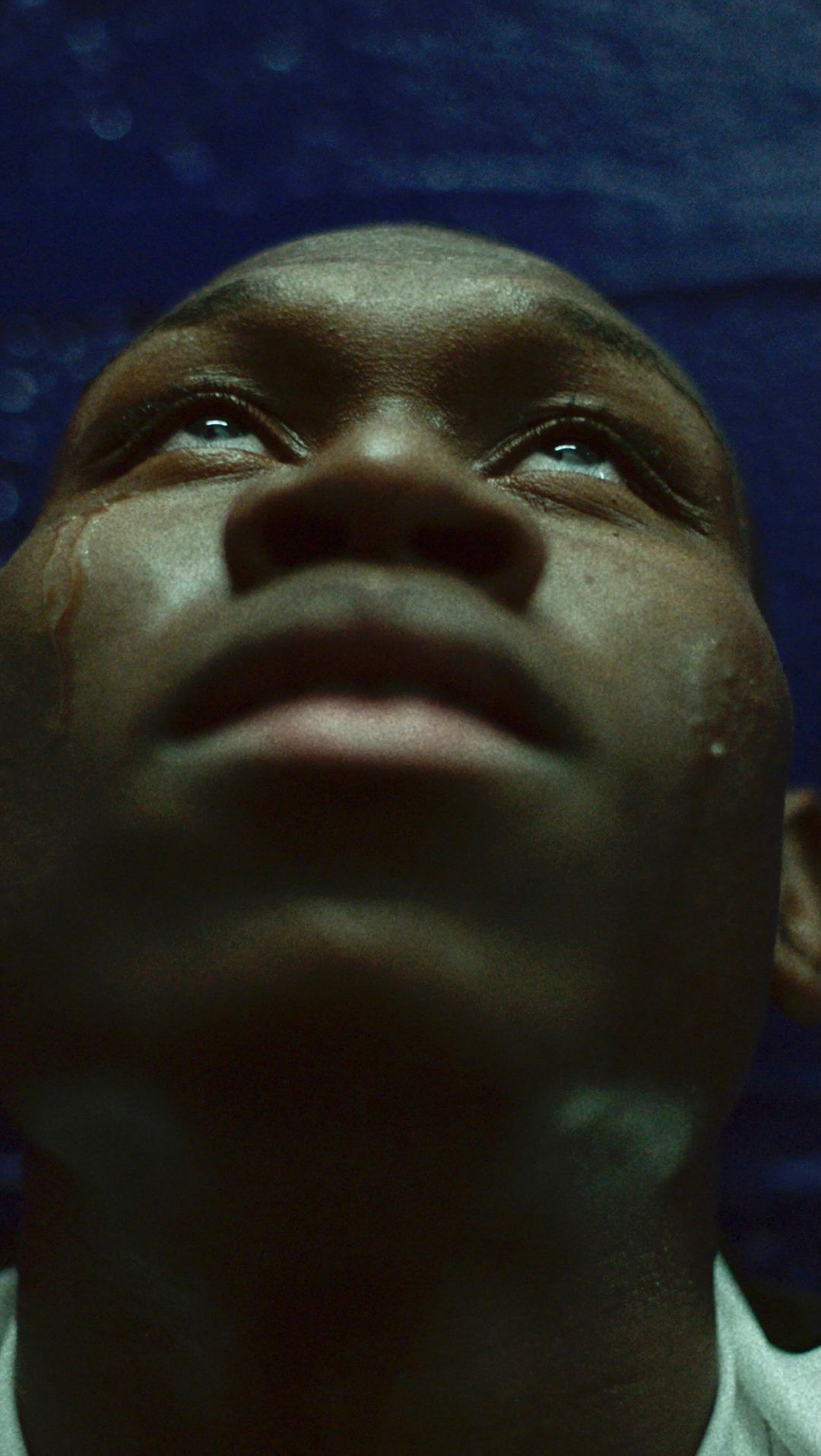 Close up portrait of a young Black man with tears streaming down his face, looking up with an emotional expression against a blue background.