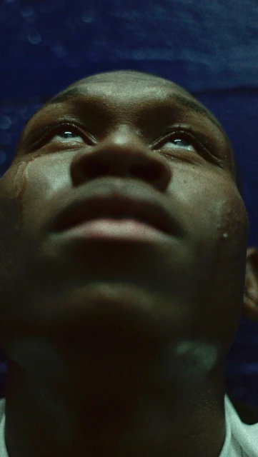 Close up portrait of a young Black man with tears streaming down his face, looking up with an emotional expression against a blue background.