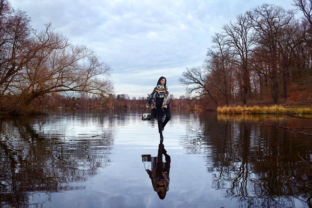 Fashionable woman stands in a lake reflecting the sky in a wide shot with fall trees on the shoreline and an overcast sky.