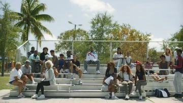 Outdoor shot of a group of diverse people sitting on bleachers under a sunny sky, near a palm tree, in sportswear and casual wear.