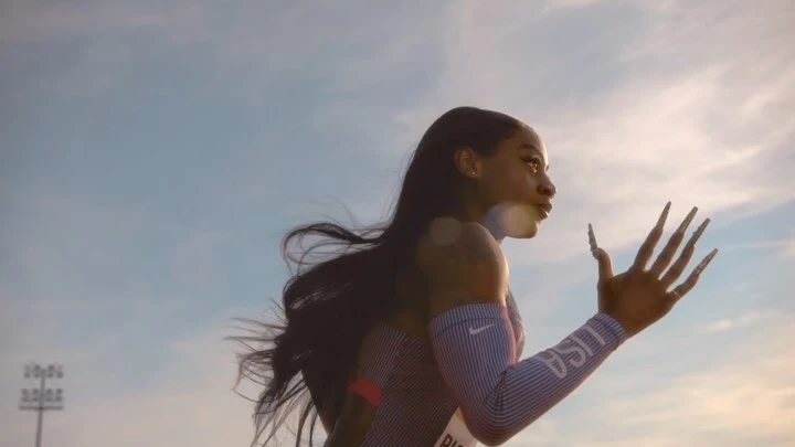 Profile shot of a woman in sportswear running, showcasing her long nails and determined expression against a partly cloudy sky.