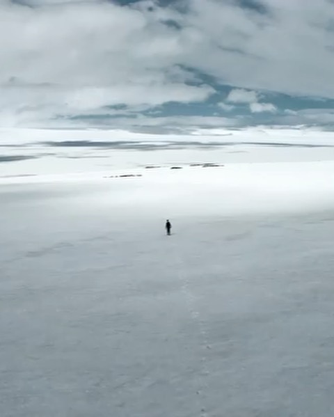 Wide landscape view of a solitary figure walking across a flat, white snow-covered plain under a cloudy sky, creating a sense of isolation.