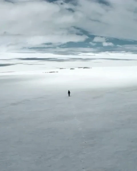 Wide landscape view of a solitary figure walking across a flat, white snow-covered plain under a cloudy sky, creating a sense of isolation.