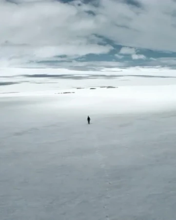Wide landscape view of a solitary figure walking across a flat, white snow-covered plain under a cloudy sky, creating a sense of isolation.