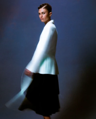 Fashionable woman in light-colored jacket and dark skirt, side pose with motion blur on a dark blue backdrop in a studio setting.