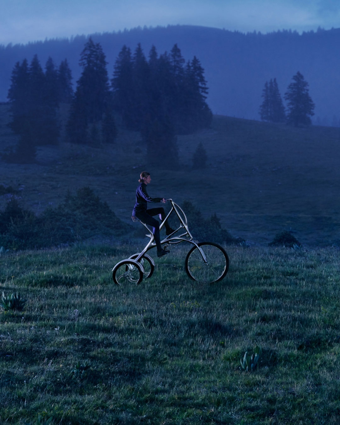 Adult riding custom tricycle through grassy meadow in hilly landscape with forest trees under an overcast blue sky.