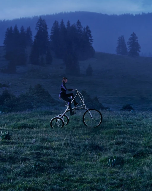 Adult riding custom tricycle through grassy meadow in hilly landscape with forest trees under an overcast blue sky.