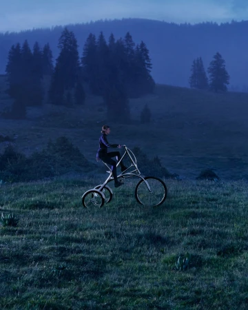 Adult riding custom tricycle through grassy meadow in hilly landscape with forest trees under an overcast blue sky.