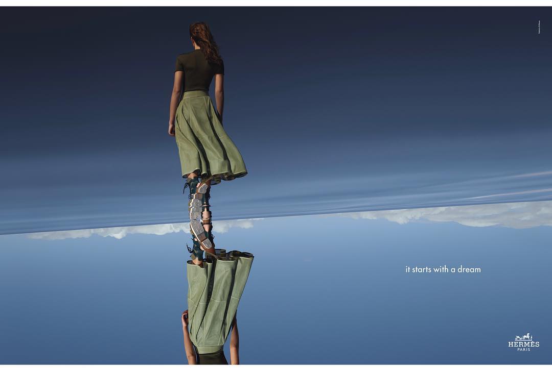 Fashion shot of a woman in olive green skirt and dark green top.  She stands on a mirrored surface reflecting the blue sky and clouds.
