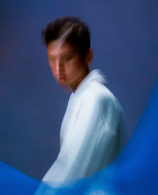 Blurred portrait of a young adult with light skin, wearing a white jacket against a blue backdrop, with a blue fabric accent.