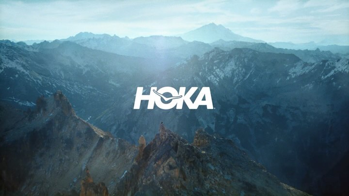 Scenic mountain range landscape with a small figure and a Hoka brand logo in white, creating a visually striking outdoor image.