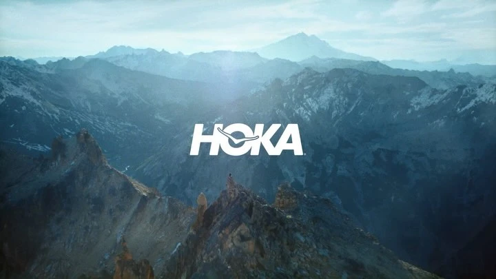 Scenic mountain range landscape with a small figure and a Hoka brand logo in white, creating a visually striking outdoor image.