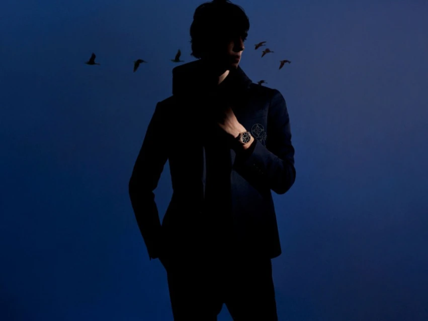 Silhouetted man in a dark jacket with birds in flight against a blue background, wearing a watch.