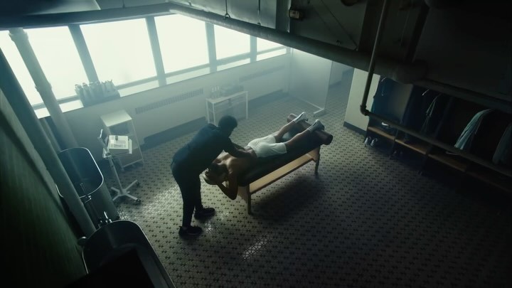 An overhead shot of two men in a locker room, one lying on a massage table as the other massages his shoulders in a bright space.